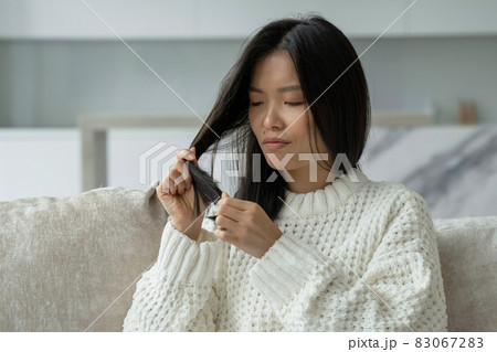 Asian woman sitting on the couch with hair problems - brittle, damaged, dry, dirty and falling out hair Asian woman sitting on the couch with hair problems - brittle, damaged, dry, dirty and falling out hair 83067283