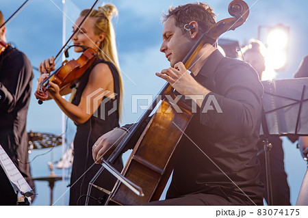 Male violoncellist playing in orchestra at outdoor concert Male violoncellist playing in orchestra at outdoor concert 83074175