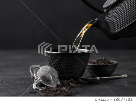 Pouring black tea from teapot to cup with strainer infuser and loose tea in ceramic bowl on black. Pouring black tea from teapot to cup with strainer infuser and loose tea in ceramic bowl on black. 83077232