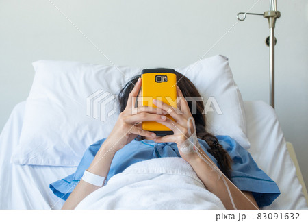 Patient woman lying on bed in hospital and receiving intravenous fluid directly into a vein but she still playing her social network on smartphone. Patient woman lying on bed in hospital and receiving intravenous fluid directly into a vein but she still playing her social network on smartphone. 83091632
