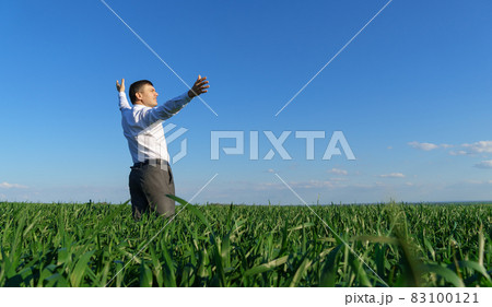 businessman poses in a field, he looks into the distance and rests, green grass and blue sky as background businessman poses in a field, he looks into the distance and rests, green grass and blue sky as background 83100121