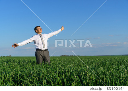 businessman poses in a field, he looks into the distance and rests, green grass and blue sky as background businessman poses in a field, he looks into the distance and rests, green grass and blue sky as background 83100164