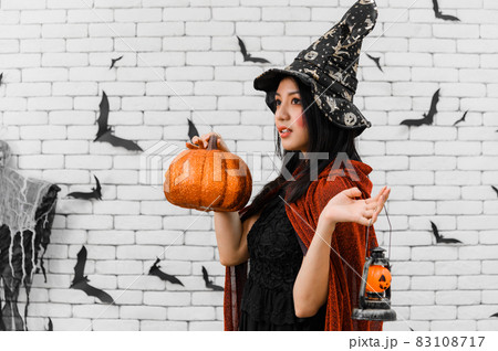 Portrait of beautiful young asian woman wearing witch costume holding pumpkin Jack O Lantern on white brick background decorated with black paper bat shape for halloween party Portrait of beautiful young asian woman wearing witch costume holding pumpkin Jack O Lantern on white brick background decorated with black paper bat shape for halloween party 83108717