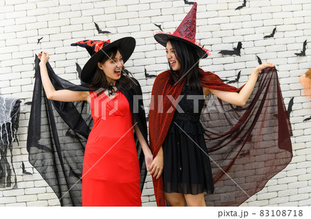 Portrait of two beautiful young asian woman in witch halloween costume on white brick background decorated with black paper bat shape for halloween party. 83108718