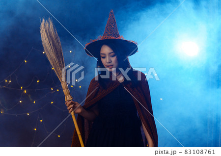 Portrait of beautiful young woman in witch halloween costume wear witches hat holding broom over spooky dark magic background - Halloween party art design. Portrait of beautiful young woman in witch halloween costume wear witches hat holding broom over spooky dark magic background - Halloween party art design. 83108761