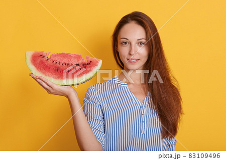 Image of young woman holding big slice of sweet watermelon while looking at camera, posing isolated over yellow background, adorable dark haired female having snack in hot summer day. People concept. 83109496