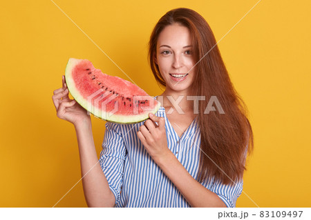 Photo of charming woman holding piece of watermelon in hands, looking directly at camera, wearing striped shirt, model posing isolated over yellow studio background. People and summertime concept. Photo of charming woman holding piece of watermelon in hands, looking directly at camera, wearing striped shirt, model posing isolated over yellow studio background. People and summertime concept. 83109497