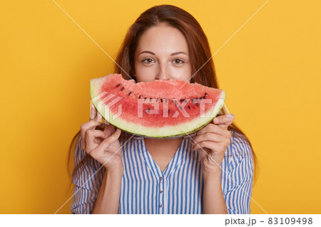 Horizontal shot of beautiful smiling young woman with slice of watermelon, female covering half of face with fruit, lady wearing blouse, having dark hair, posing isolated over yellow background. 83109498