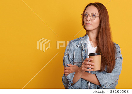 Close up portrait of pensive woman with dark hair, looking aside, contemplates about something, wearing white t shirt, denim jacket abd eyewear, holding disposable cup of coffee, over yellow wall. Close up portrait of pensive woman with dark hair, looking aside, contemplates about something, wearing white t shirt, denim jacket abd eyewear, holding disposable cup of coffee, over yellow wall. 83109508