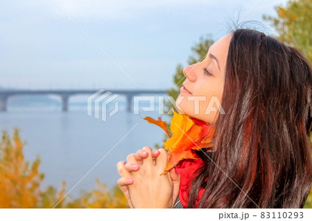 Pretty brunette girl holding yellow autumn leaves standing on a river shore. Pretty brunette girl holding yellow autumn leaves standing on a river shore. 83110293