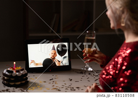 Online birthday. Holiday greeting. Virtual party. Unrecognizable woman holding glass champagne celebrating holiday cake with festive man on laptop dark home interior. Online birthday. Holiday greeting. Virtual party. Unrecognizable woman holding glass champagne celebrating holiday cake with festive man on laptop dark home interior. 83110508
