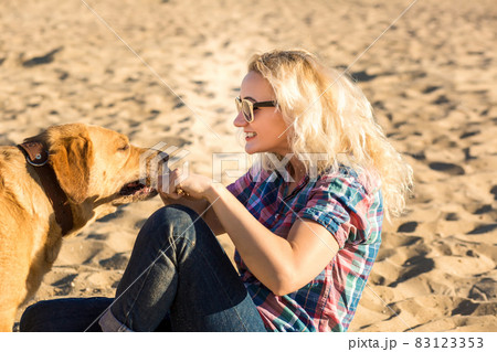 Portrait of young beautiful woman in sunglasses sitting on sand beach hugging golden retriever dog. Girl with dog by sea. Happiness and friendship. 83123353