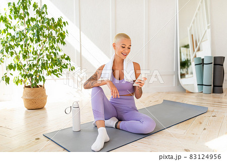 Active lifestyle concept. Young lady relaxing on yoga mat with smartphone after her home workout Active lifestyle concept. Young lady relaxing on yoga mat with smartphone after her home workout 83124956