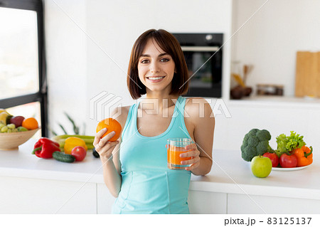Young woman drinking juice and holding orange, standing near kitchen table full of fruits and smiling at camera 83125137
