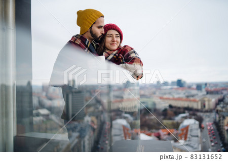 Young couple wrapped in blanket standing and hugging outdoors on balcony, shot through glass. 83131652