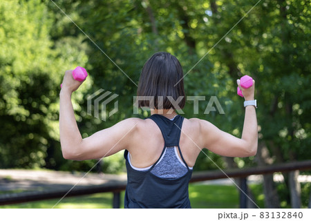 Fitness trainer conducts workout in the park. Girl holds small dumbbells and does the exercise. Back view. Fitness trainer conducts workout in the park. Girl holds small dumbbells and does the exercise. Back view. 83132840