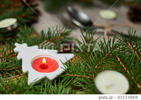 Burning candle in a Christmas tree on a served Christmas table against a background of fir branches and cones 83133069