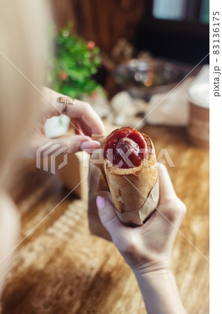 Close-up of a hot dog in hand. The girl has a French manicure. There is a lot of ketchup on top and sauce on the food. The woman wrapped her hands around the bun, creating a hint of intimacy. 83136175