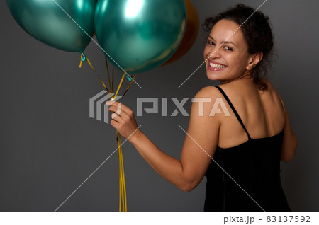 Joyful charming woman smiles with beautiful smile, looks at camera through her shoulder and holds green metallic shiny inflated balloons. Birthday event, Christmas, New Year concept with copy space Joyful charming woman smiles with beautiful smile, looks at camera through her shoulder and holds green metallic shiny inflated balloons. Birthday event, Christmas, New Year concept with copy space 83137592