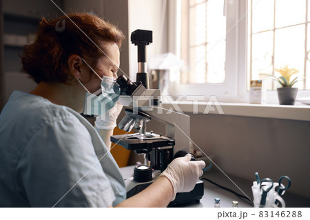 Mature lab assistant in mask looks into microscope researching sample at workplace Mature lab assistant in mask looks into microscope researching sample at workplace 83146288