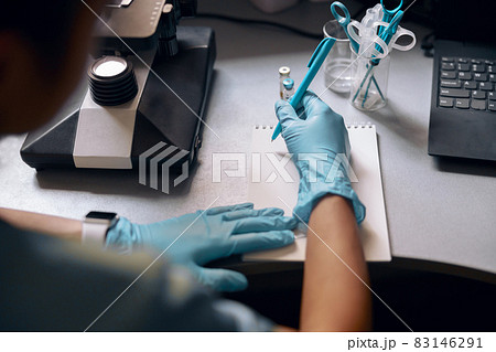 Woman in gloves writes in notebook near microscope at table in laboratory Woman in gloves writes in notebook near microscope at table in laboratory 83146291