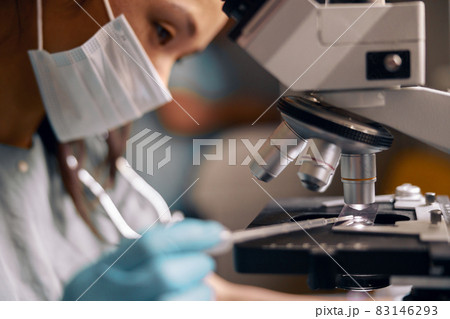Asian technician with mask drips liquid sample onto slide working with microscope in clinic 83146293