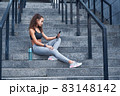 Young athletic woman in sport clothes sitting on stairs of city stadium with phone in hands posting her training results 83148142