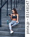 Young athletic woman in sport clothes sitting on stairs of city stadium with phone in hands posting her training results 83148147