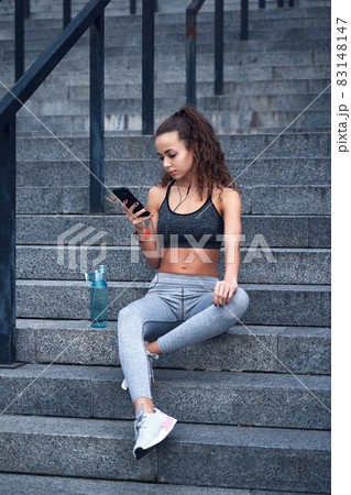 Young athletic woman in sport clothes sitting on stairs of city stadium with phone in hands posting her training results 83148147