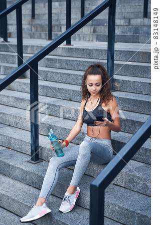 Young athletic woman in sport clothes sitting on stairs of city stadium with phone in hands posting her training results 83148149