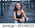 Young athletic woman in sport clothes smiling and listening to music while sitting on stairs of city stadium 83148150