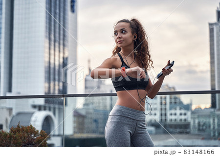 Young athletic woman checking her training results on sport watch standing with phone and listening to music 83148166