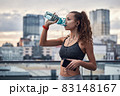 Side shot of young athletic woman drinking water from sport bottle while listening to music on city background 83148167