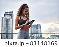 Young athletic woman in sport clothes standing on city background with phone in hands posting her training results 83148169