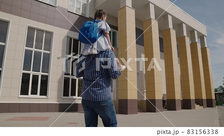 Dad carries on his shoulders little kid with backpack to school, girl with school bag rides her father around the schoolyard, happy family an elementary school, first grader with parent goes to class Dad carries on his shoulders little kid with backpack to school, girl with school bag rides her father around the schoolyard, happy family an elementary school, first grader with parent goes to class 83156338