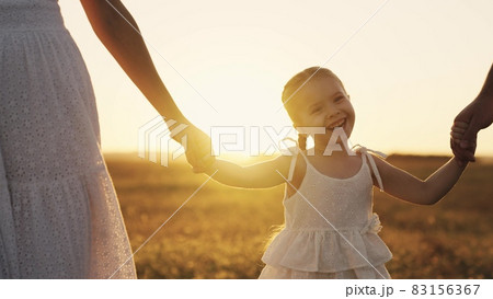 happy baby smiling at sunset with parents, mother, father and child walk in the sun, hold kid by hands, young family, taking care of their little daughter, family walk in evening park in orange light 83156367