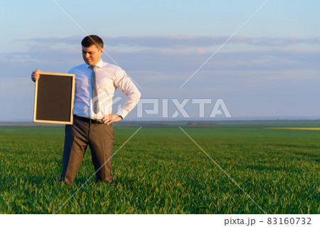 businessman holds a blackboard with an empty space and poses on a green grass field - business concept businessman holds a blackboard with an empty space and poses on a green grass field - business concept 83160732