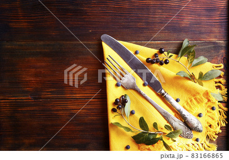 Thanksgiving table setting concept. Fork and knife on yellow textile napkin with green leaves & berries on brown wooden table. November mood background. Copy space, close up, top view, flat lay. Thanksgiving table setting concept. Fork and knife on yellow textile napkin with green leaves & berries on brown wooden table. November mood background. Copy space, close up, top view, flat lay. 83166865