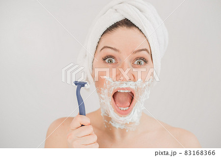 Funny portrait of a woman with shaving foam on her face holding a razor on a white background. The girl removes the mustache and beard Funny portrait of a woman with shaving foam on her face holding a razor on a white background. The girl removes the mustache and beard 83168366