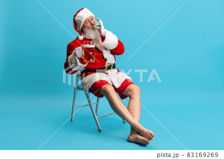 Full-length portrait of senior man wearing Santa Claus costume, holding glass of lager foamy beer isolated over blue background 83169269