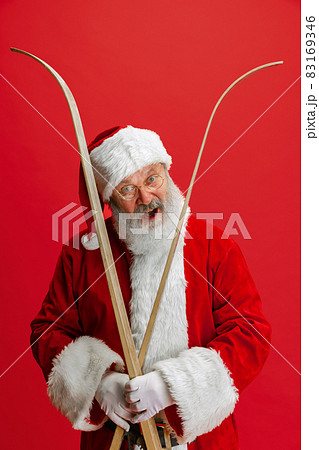Cropped portrait of joyful senior man wearing Santa Claus costume, holding skis isolated over red background. Winter holiday season Cropped portrait of joyful senior man wearing Santa Claus costume, holding skis isolated over red background. Winter holiday season 83169346