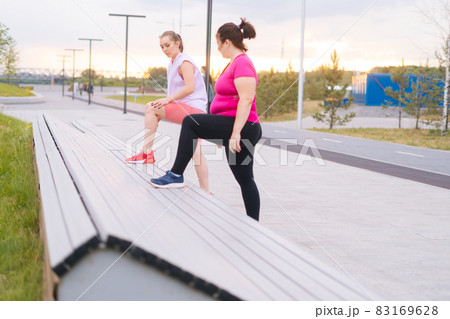 Overweight young woman doing single leg squats using bench with personal trainer outdoor in summer morning. 83169628