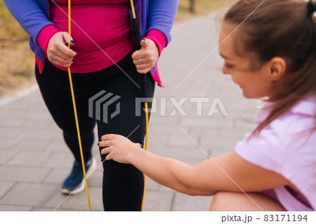 Close-up cropped shot obese overweight young woman doing exercises using fitness tape for weight loss with personal trainer outdoor in summer day. 83171194