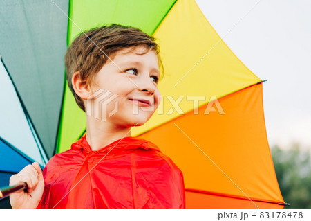 Portrait of a laughing school boy with rainbow umbrella behind. Smiling kid holds colourful umbrella on his shoulder. Cheerful child in a red raincoat holding multicolor umbrella 83178478