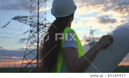 A woman power engineer with a project of new electrical works, an electrician walks against the background of high towers of power plants, the expansion of electrical equipment of voltage volts A woman power engineer with a project of new electrical works, an electrician walks against the background of high towers of power plants, the expansion of electrical equipment of voltage volts 83184457