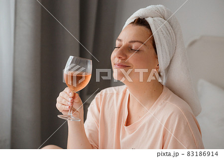 Beautiful happy young woman after shower with glass of wine in bedroom. Staying at home in isolation during quarantine lockdown. Concept of celebrating birthday in bed alone Beautiful happy young woman after shower with glass of wine in bedroom. Staying at home in isolation during quarantine lockdown. Concept of celebrating birthday in bed alone 83186914
