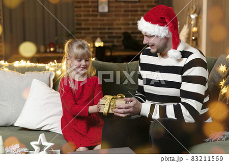Merry Christmas and Happy New Year. Christmas gift surprise. A little happy smiling daughter girl opens a present. Father holding and giving giftbox indoors in house with festive Christmas decorations 83211569