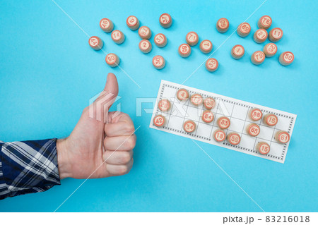 Man shows sign like. Wooden barrels with numbers and a card for playing lotto or bingo. Board game. Gambling game with random element. Blue background. Man shows sign like. Wooden barrels with numbers and a card for playing lotto or bingo. Board game. Gambling game with random element. Blue background. 83216018