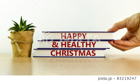 Books with text 'happy and healthy christmas' on beautiful wooden table, white background. Male hand and house plant. Business and new year concept. Copy space. 83219247