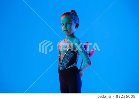 Portrait of little girl, professional rhythmic gymnast performing with gymnastic clubs isolated over blue background in neon Portrait of little girl, professional rhythmic gymnast performing with gymnastic clubs isolated over blue background in neon 83226009